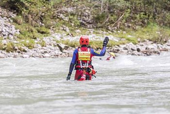 Ein Strömungsretter von hinten steht in rot-blauer Einsatzkleidung im fließenden Gewässer und hebt rechte flache Hand